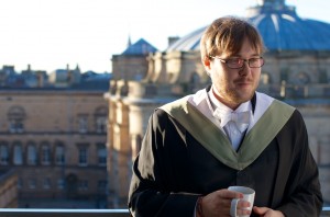 Young man with beard and graduation gown drinking tea